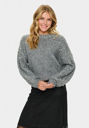 Maglione grigio lavorato a maglia con dettagli in perle sulle maniche e orlo a costine. Presenta un colletto rotondo e un design leggermente accorciato. Abbinato a una gonna nera.