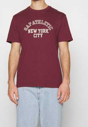 Bordeauxfarbene Baumwoll-T-Shirt mit Rundhalsausschnitt, kurzen Ärmeln und dem weißen Druck "GAP ATHLETIC NEW YORK CITY" über der Brust. Kombiniert mit hellen Jeans.