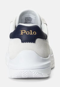 Vit sneaker med marinblå läderdetalj vid hälen, med ett guld "Polo"-logotyp, texturerade mockadetaljer och gummisula med ett mönster av däckspår.