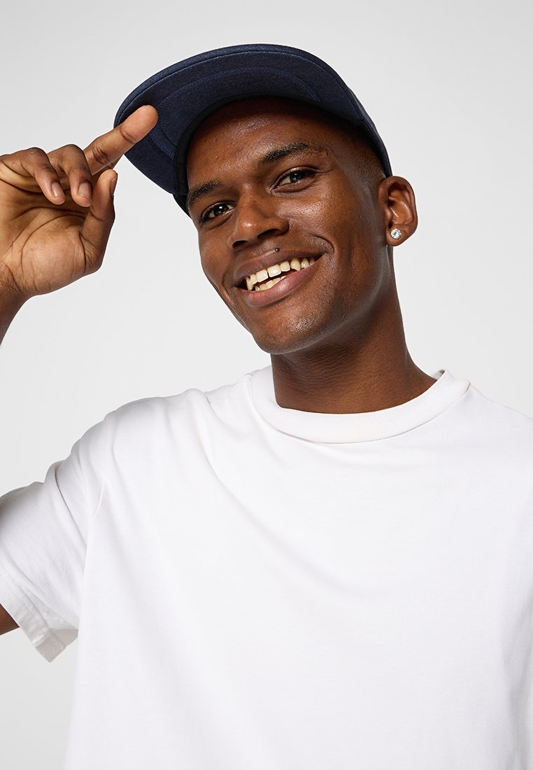 Hombre sonriente con una camiseta blanca y una gorra oscura, tocando el borde de la gorra con su mano derecha frente a un fondo claro y liso.