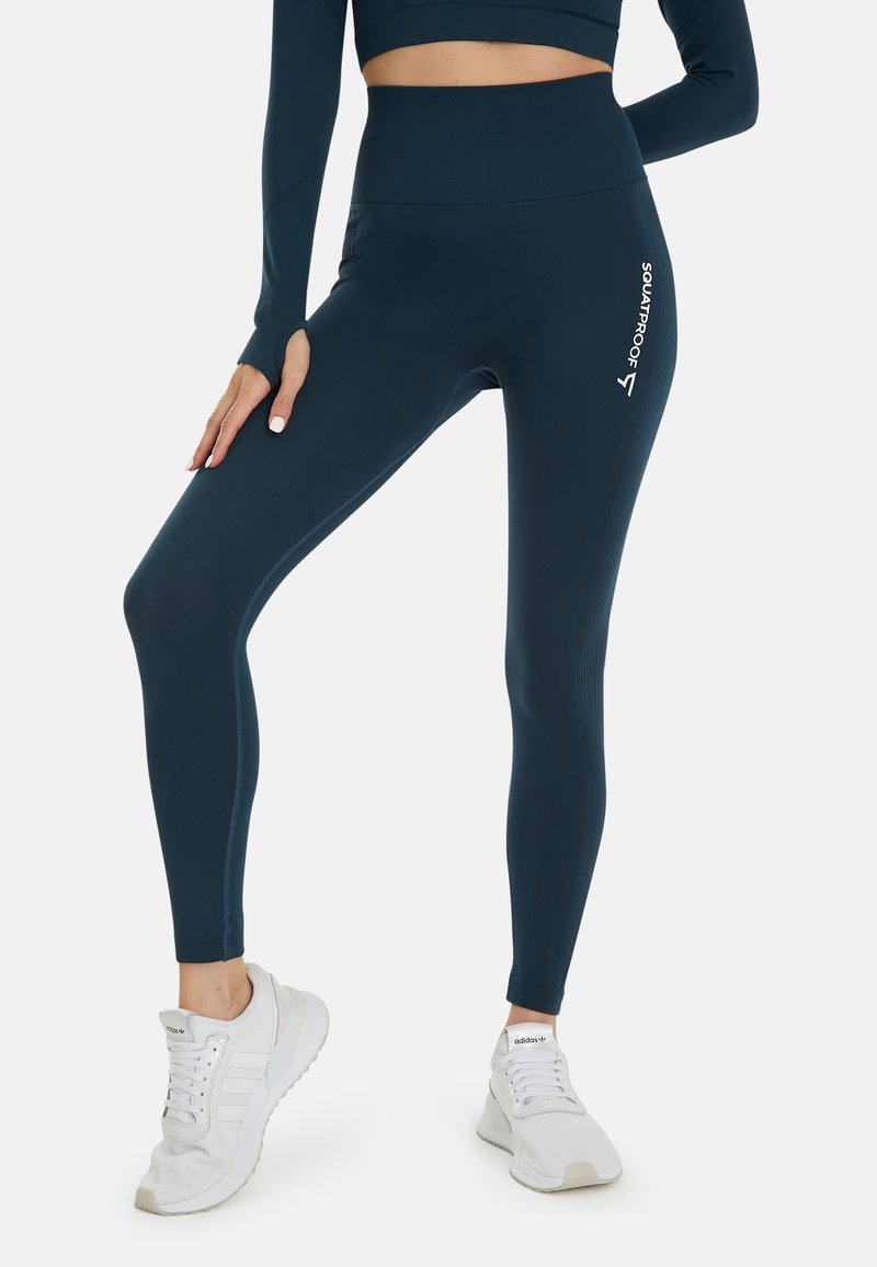 Tmavě modré legíny s vysokým pasem a ribovanou texturou s logem "SQUATPROOF". V kombinaci s bílými sportovními botami.