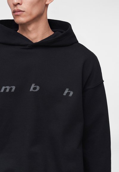 Jeune personne portant un sweat à capuche noir avec des lettres grises "m b h" espacées sur la poitrine sur un fond uni.