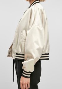 Veste bomber en satin crème avec poignets et ourlet côtelés à rayures noires, dotée d'une fermeture à boutons-pression et de poches latérales. Texture lisse.