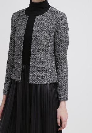 Kobieta ubrana w czarny golf, czarną plisowaną spódnicę i teksturowaną czarno-białą otwartą na przodzie kurtkę, stojąca na tle o neutralnym kolorze.