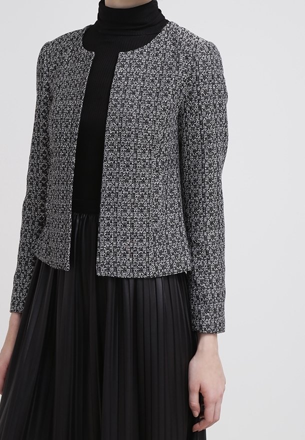 Femme portant un col roulé noir, une jupe plissée noire et une veste noire et blanche texturée ouverte à l'avant, debout devant un fond neutre.