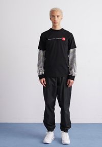 Zwarte T-shirt met witte tekst en rood logo, gelaagd over zwart-wit gestreepte lange mouwen, gecombineerd met losse, zwarte, waterdichte broek en witte sneakers.