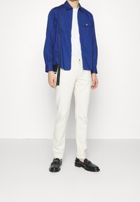 Camisa azul de manga larga, pantalones blancos ajustados, mocasines negros. Cuenta con un bolsillo en la camisa y una pequeña bolsa negra con una correa al hombro.