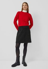 Frau, die einen roten Pullover über einem weißen Rollkragenpulli, einen schwarzen glitzernden A-Linien-Rock, schwarze Strumpfhosen und schwarze Stiefeletten trägt, steht mit den Händen hinter dem Rücken.