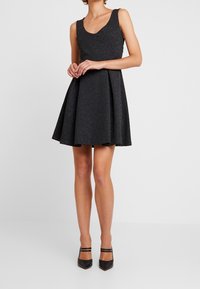 Svart ärmlös klänning med rundad halsringning, utsvängd kjol och texturerad tyg. Bärs med svarta högklackade skor med ankelsnören.