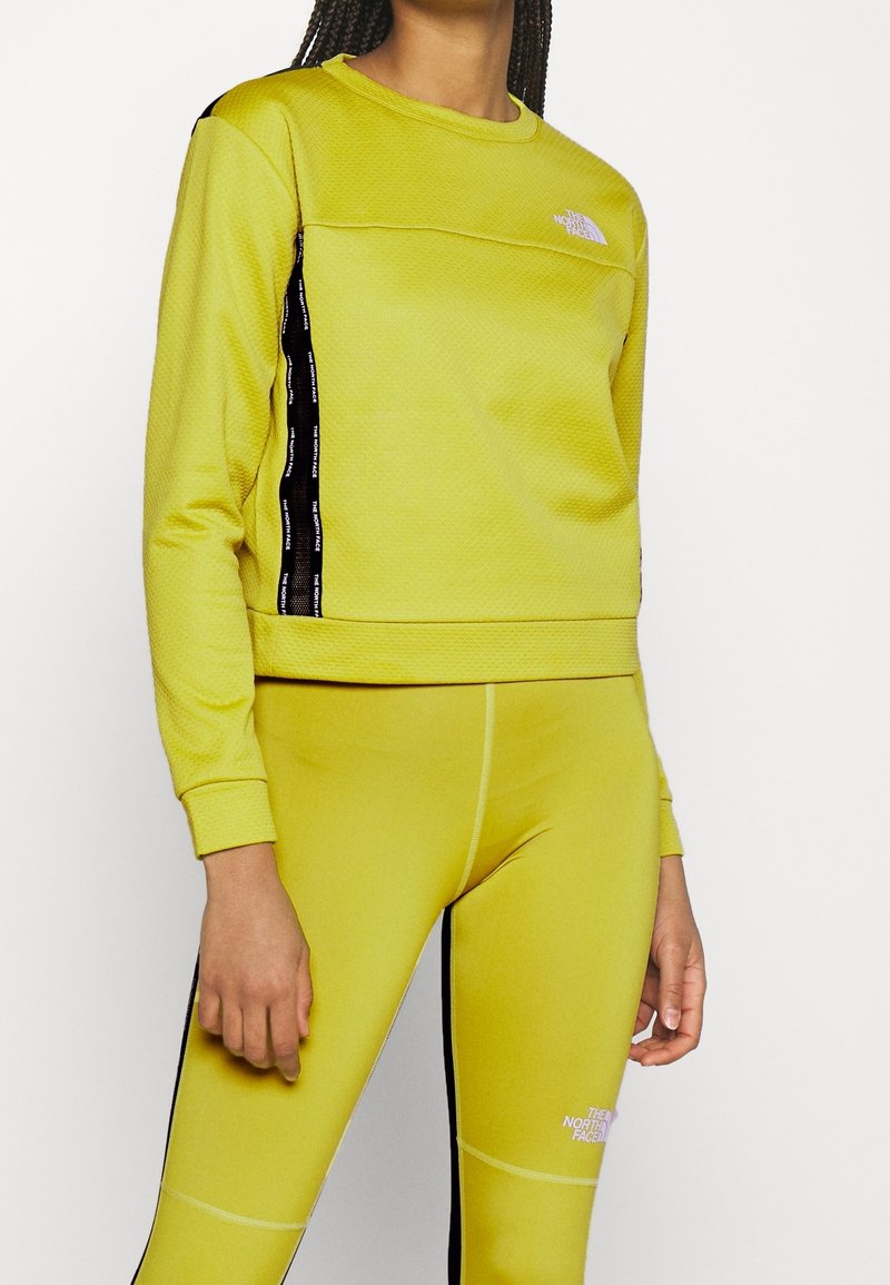 Femme portant un haut à manches longues jaune et des leggings assortis avec des bandes latérales noires et les logos The North Face.