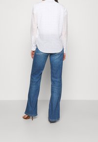 Blouse blanche avec un motif bleu subtil, manches longues et poignets boutonnés, associée à un jean flare bleu clair et des sandales à talons beiges.