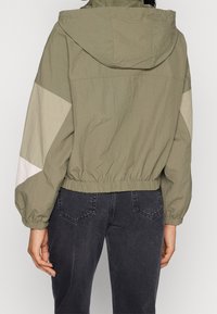 Person wearing cropped olive green hooded jacket with beige color-blocked sleeves and black high-waisted jeans, shown from the back.
