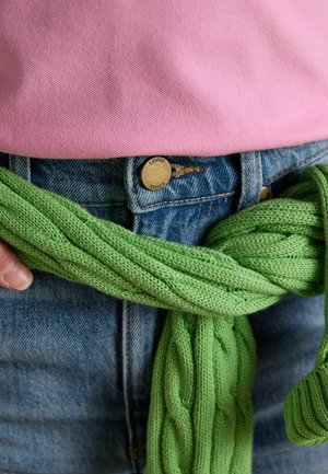 Henkilö, jolla on siniset farkut, vyötärölle sidottu vihreä neulepusero ja sisään tungettu vaaleanpunainen tekstuuroitu paita.