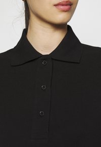 Nahaufnahme einer Person, die ein schwarzes, geknöpftes Poloshirt trägt, wobei ein Teil des Halses, der Lippen und des Kragenbereichs sichtbar ist.