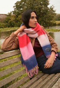 Viacfarebná pletená šatka s ružovými, červenými a modrými pruhmi, ozdobená strapcami na koncoch a kruhovou látkovou nášivkou. Mäkká textúra.