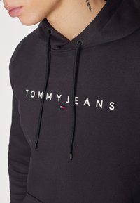 Čierna mikina s kapucňou vyrobená z bavlny s predným vreckom, ktorá má biele logo "TOMMYJEANS" a malý viacfarebný prízvuk na boku.