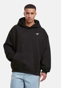 Sudadera con capucha negra, con un bolsillo frontal, textura suave y pequeño logo blanco en el pecho. Combinada con jeans de color azul claro.