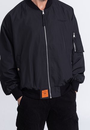 Mand iført sort bomberjakke med frontlynlås, lynlåslomme på ærmet, ribstrikkede manchetter og orange mærkat nær kanten, hænderne i bukselommerne.