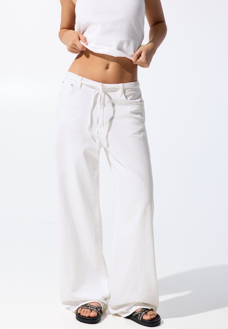 Witte wijde broek van lichtgewicht stof, met een trekkoord in de taille en een subtiele textuur. Gepaard met zwarte strap-sandalen.