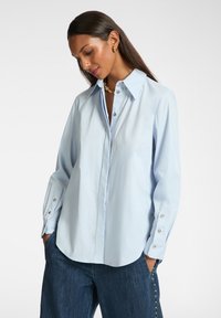 Camisa azul clara de botones con un cuello estructurado, mangas largas con acentos de botones plateados y un corte relajado. Tejido suave y tejido.