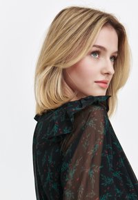 Black blouse with floral pattern featuring green leaves and red berries. Sheer fabric with ruffled collar detail. Hair styled in soft waves.
