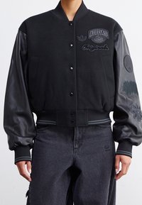 Zwarte varsityjack met een wollen lichaam en leren mouwen, voorzien van verschillende geborduurde logo's en een geribbelde zoom en manchetten.