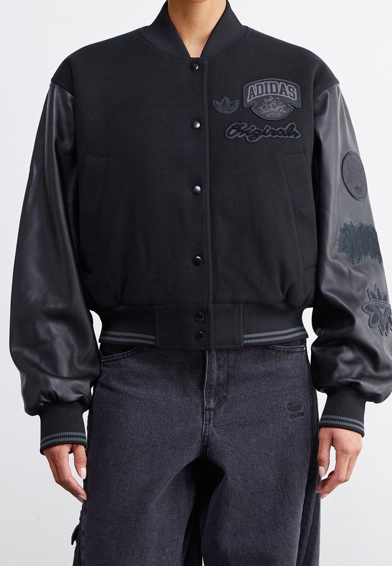 Zwarte varsityjack met een wollen lichaam en leren mouwen, voorzien van verschillende geborduurde logo's en een geribbelde zoom en manchetten.
