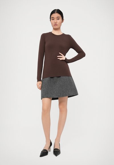 Bruine lange mouwen top gecombineerd met een grijze flared rok, met een gekorrelde afwerking. Model draagt puntige zwarte hakken. Eenvoudige achtergrond.