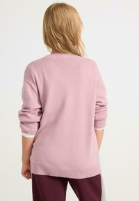 Ljusrosa tröja med ribbat krage och mudd, med en lös design och mjuk textur, i kombination med mörka byxor. Vy från baksidan.