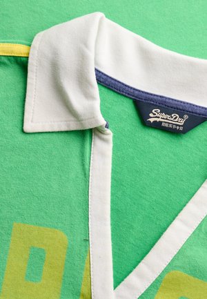 Zielona koszulka polo z białym kołnierzem, niebieską wewnętrzną lamówką kołnierza oraz częściowo widocznym żółtym nadrukiem, z widoczną etykietą marki SuperDry.