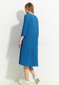 Blaues, strukturiertes Midikleid mit drei Viertelärmeln und lockerer Passform, kombiniert mit weißen Sneakern mit einem hellgrünen Logo.