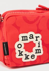 Κόκκινη υφασμάτινη θήκη με ένα κυκλικό λευκό ταμπελάκι που γράφει «marimekko» με μαύρα γράμματα και μια μαύρη ετικέτα Marimekko στο πλάι.