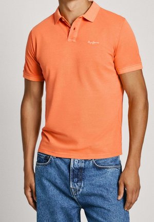 Mężczyzna ubrany w pomarańską krótką koszulkę polo i niebieskie dżinsy na jednolitym tle.