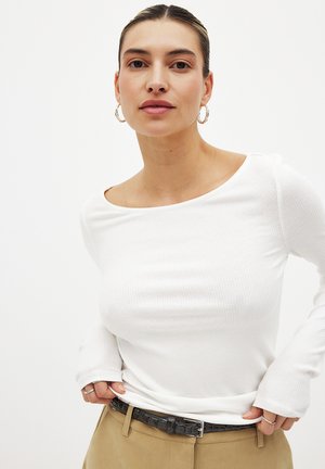 White ribbed long-sleeve top with a wide neckline, paired with beige trousers and a textured black belt. Metal hoop earrings are visible.