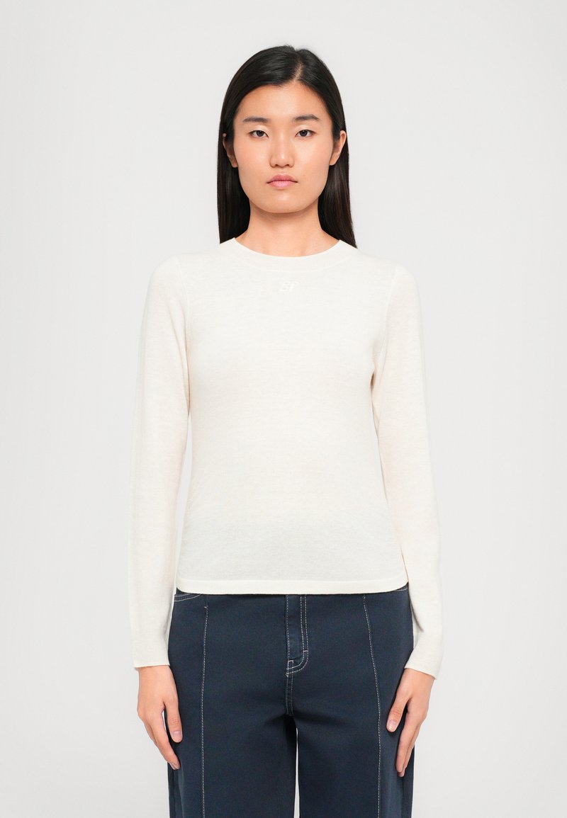 Kreemikaskine pikkade varrukatega topp, mis on valmistatud pehmest ja kergest kangast. Omab ümmargust kaelust ja väikest, diskreetset logo rinnal.