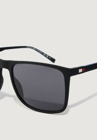 Mustat suorakulmaiset aurinkolasit tummilla linsseillä ja pienellä punavalkosinisellä logolla sankojen kohdalla vaalealla taustalla.