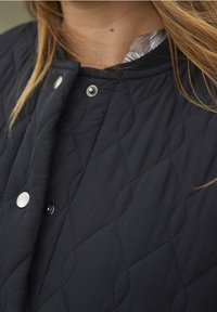 Musta tikattu takki timanttikuvioilla, jossa hopeanväriset painonapit ja kontrastivärinen ruudullinen vuori. Pehmeä tekstuuri.