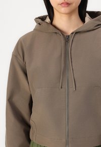 Chaqueta corta con capucha en verde oliva, fabricada con un tejido suave, que presenta un cierre frontal con cremallera, un gran bolsillo y detalles de costura sutiles.