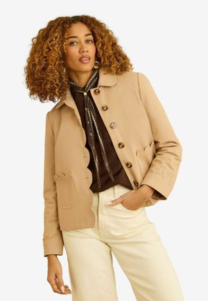 Vrouw met krullend haar die een beige jas met gekartelde rand, een bruin topje, een patroon sjaal en lichtgekleurde broek draagt, poseert met hand in de zak.