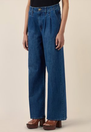 Frau trägt hoch taillierte, weit geschnittene blaue Jeans mit Falten, kombiniert mit braunen Plateau-Open-Toe-Pumps, steht vor einem einfarbigen Hintergrund.