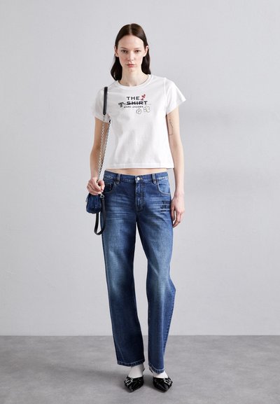 Hvit avkortet t-skjorte med grafisk tekst, parret med blå vide jeans. Modellen holder en liten svart crossover-veske med kjedeakse.