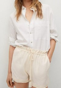 Chemise blanche en lin à boutons avec une poche, associée à un short crème avec une taille élastique et un cordon de serrage.