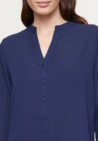 Blouse marine avec un décolleté en V, des manches longues, une fermeture à boutons à l'avant, et une texture lisse et douce. Aucun motif ni accent supplémentaire visible.