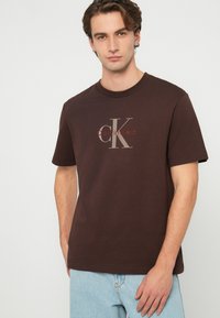 T-shirt castanha de algodão com um grande logótipo creme da Calvin Klein na parte da frente, complementada por mangas curtas e decote redondo.