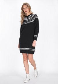 Abito nero lavorato a maglia con motivo geometrico bianco attorno al collo e all'orlo, dotato di maniche lunghe e vestibilità comoda. Indossato con sneakers bianche.