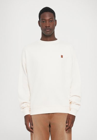 Man standing wearing a plain off-white sweatshirt with a small brown logo on the chest and light brown pants against a white background.