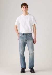 Young man standing wearing a plain white t-shirt, light blue ripped jeans, and black leather shoes against a plain background.