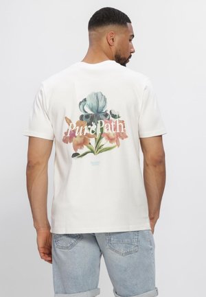 Mann trägt weißes T-Shirt mit buntem Blumenmuster und dem Schriftzug „Pure Path“ auf dem Rücken, kombiniert mit hellen Jeansshorts.