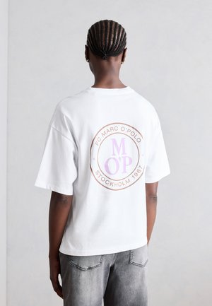 Personne aux cheveux tressés portant un t-shirt blanc avec un logo circulaire "FC Marc O'Polo Stockholm 1967" dans le dos et un jean gris délavé.