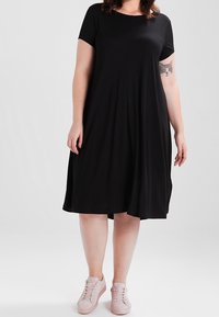 Czarna sukienka z krótkim rękawem, okrągłym dekoltem, zwiewnym krojem i długości do kolan, w zestawieniu z jasnoróżowymi sneakersami. Gładka tekstura materiału.